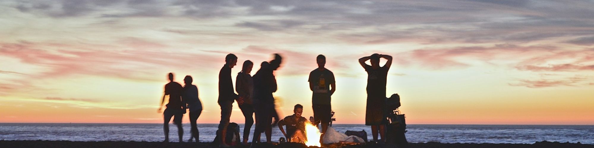 A group of people gathered around a bonfire on the beach during sunset, with the ocean and colorful sky in the background.