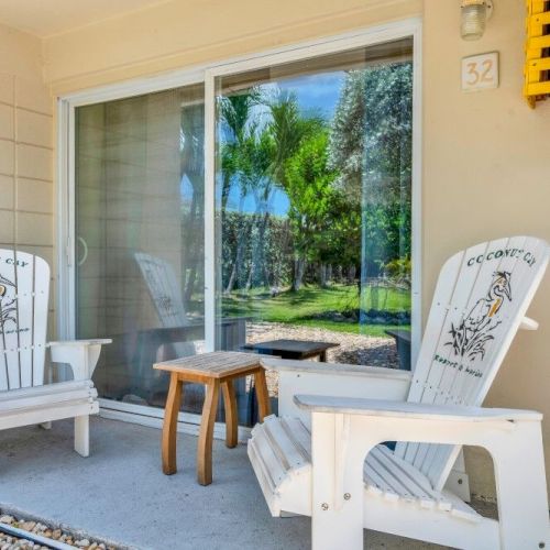 Two white chairs and a small wooden table sit on a patio with a glass door, overlooking a yard with trees.