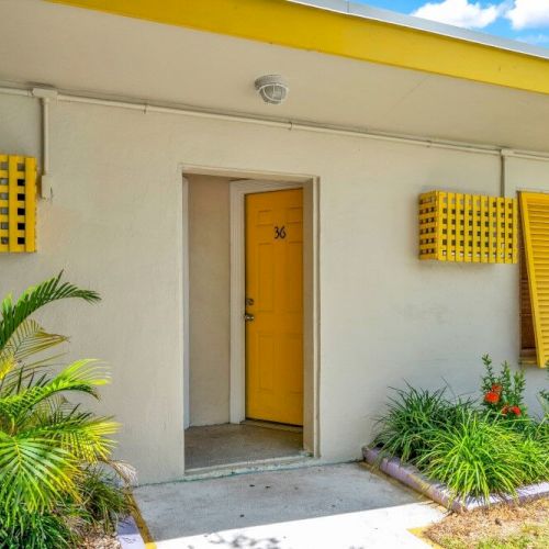 A single-story building with a yellow door and shutters, surrounded by plants, under a blue sky with some clouds.