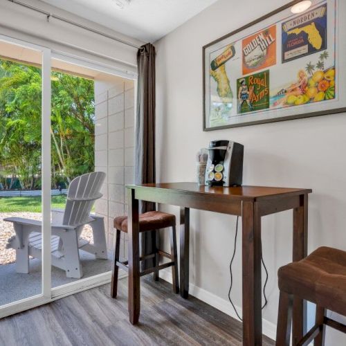 A small indoor coffee area with a table, stools, and a coffee machine. A large window shows a patio with a chair and greenery outside.