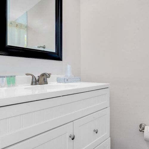 The image shows a bathroom sink with a mirror, toiletries, and a towel holder on the wall with a roll of toilet paper.