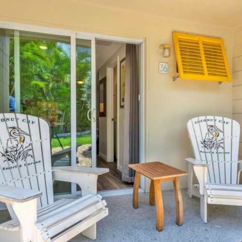 Two white chairs and a small wooden table on a patio, with a sliding glass door and green foliage in the background.