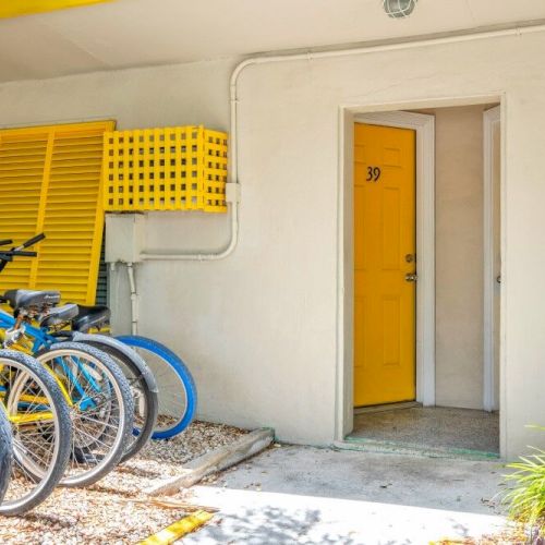 The image shows parked bicycles with blue wheels and a yellow door numbered 39, set against a beige wall with yellow accents and greenery nearby.