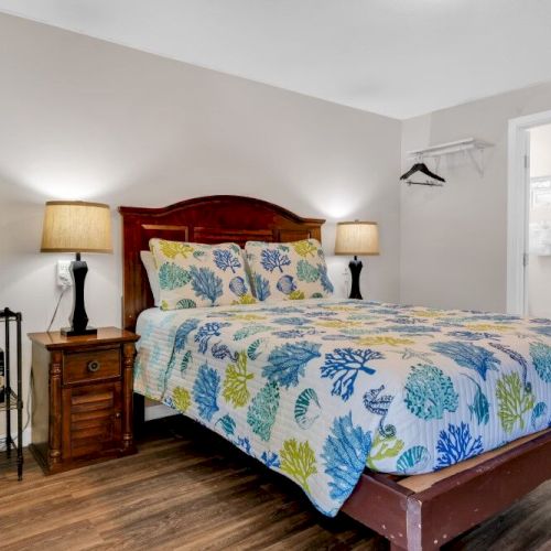 A cozy bedroom with a patterned bedspread, two lamps, a mini fridge, and a small fireplace. There's an open door to a bathroom.