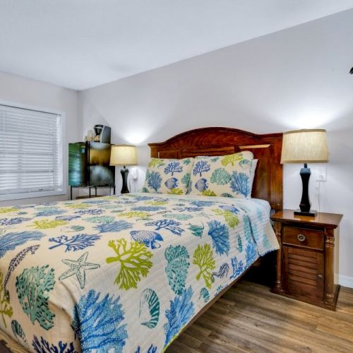 A cozy bedroom with a bed featuring a colorful floral bedspread, two lamps on wooden nightstands, and a window with blinds.