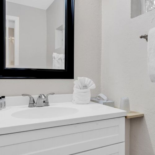 A bathroom with a sink, mirror, toiletries, towel with "ENJOY" label, and a cup on a shelf. Minimalist and clean design.