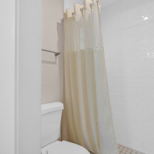 The image shows a bathroom with a white toilet, a shower with a white curtain, and tiled walls.