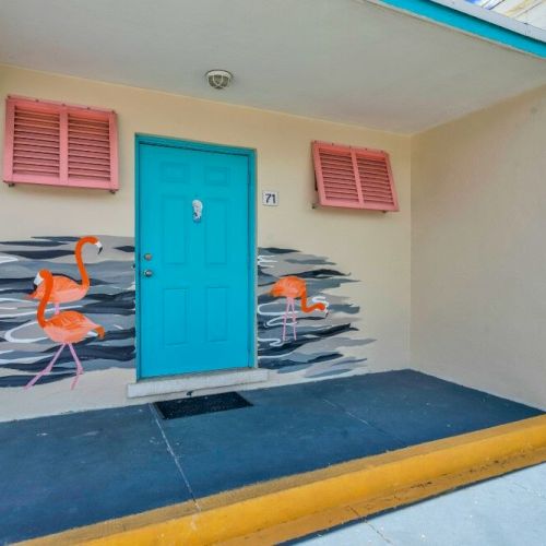 A vibrant turquoise door with flamingo murals and colorful shutters is featured, set against a beige wall under a covered entrance.