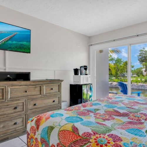 A brightly decorated bedroom with a dresser and TV; a window shows a view of greenery and a patio with colorful chairs outside.