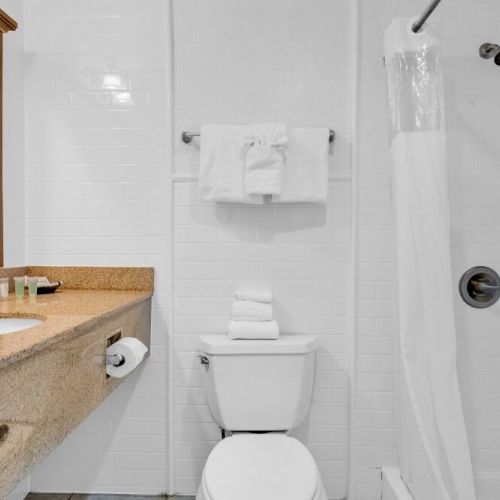 The image shows a clean, white bathroom with a sink, countertop, toilet, towels, and a shower with a curtain.