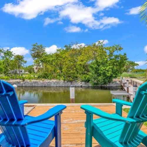 The image shows two blue Adirondack chairs on a wooden deck facing a serene canal surrounded by lush greenery under a partly cloudy sky.