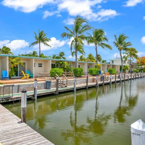 A row of houses with palm trees, colorful chairs, and a dock alongside a calm waterfront under a bright blue sky with clouds.