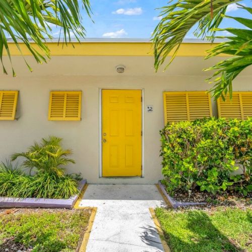 A quaint building with a yellow door and shutters is surrounded by lush greenery and palm leaves under a clear blue sky.