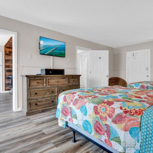 A bedroom with colorful bedding, a wooden dresser, a wall-mounted TV, and three doors leading to other rooms. Bright and neatly arranged.