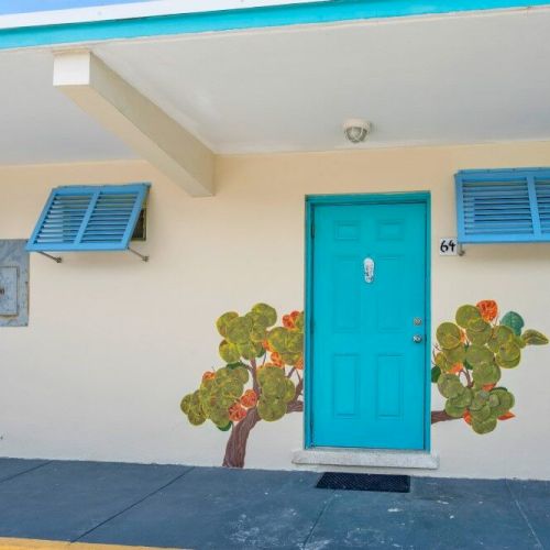 A building facade with a turquoise door and colorful shutters. Decorative mural of a tree by the door, creating a cheerful vibe.