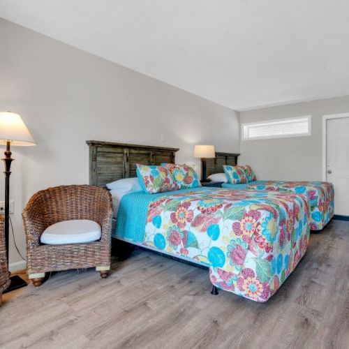 A cozy bedroom with two beds, vibrant floral bedding, wicker chairs, a floor lamp, and a small kitchenette area.