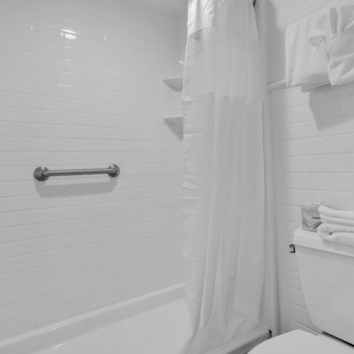 The image shows a clean, white-tiled bathroom with a bathtub, shower curtain, and towels neatly stacked on a toilet tank lid.