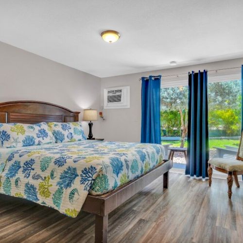 A cozy bedroom with a floral bedspread, wooden furniture, blue curtains, and a view of a garden through the windowed door.