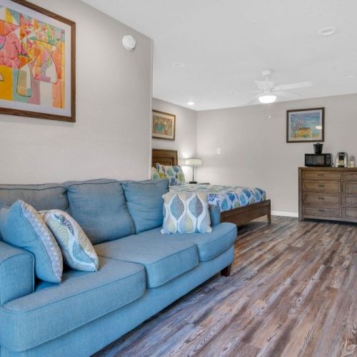 The image shows a cozy room with a blue sofa, a bed, abstract artwork, a dresser, and a wall-mounted TV, with wood flooring throughout.