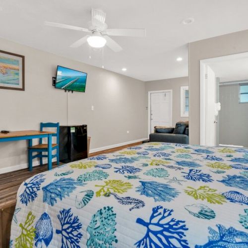 A room with a bed featuring a colorful ocean-themed cover, a small table with chairs, a TV mounted on the wall, and a doorway to a bathroom.