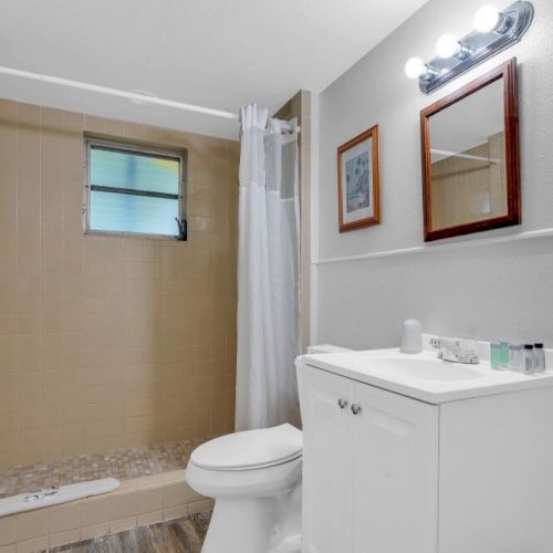 A bathroom with a shower, toilet, and sink. Towels are hung, and toiletries are on the sink. A mirror and a wall light are above.