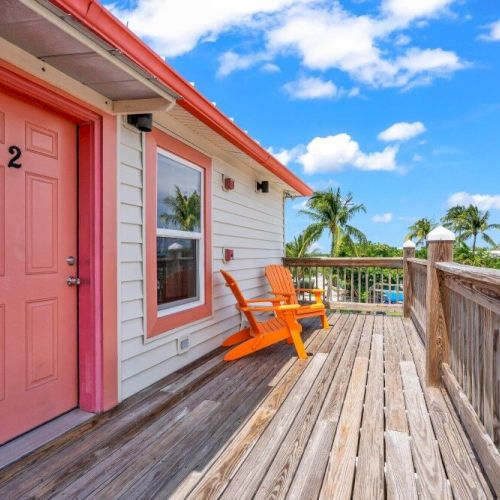 A pink door with the number 2, orange chairs on a wooden deck, and palm trees under a blue sky with clouds.