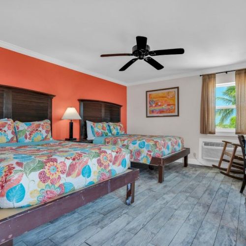 The image shows a colorful hotel room with two beds, floral bedding, an orange accent wall, a ceiling fan, and a window with a pleasant view.