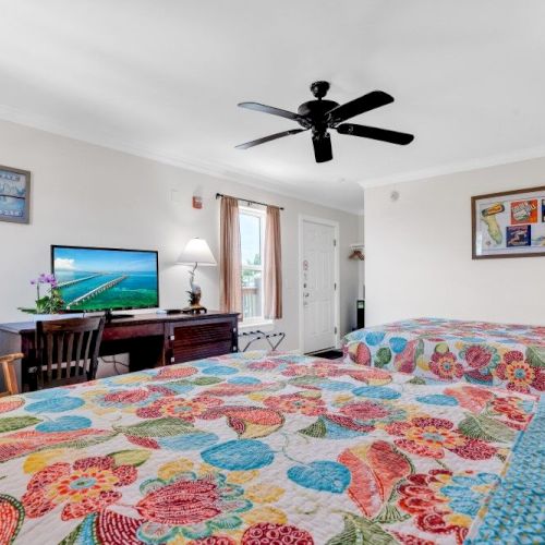 A cozy room with two colorful beds, a TV, a chair, a fan, and decor. The room has a window and a door leading outside.