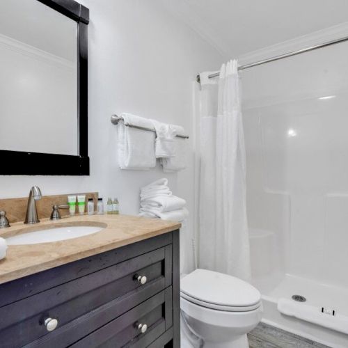 A modern bathroom with a sink, mirror, towels, and toiletries on a counter, alongside a toilet and a white shower with a curtain.