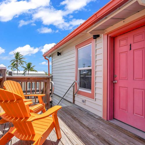 A porch with two orange chairs, a pink door, and a view of palm trees under a blue sky with clouds, creating a tropical vibe.