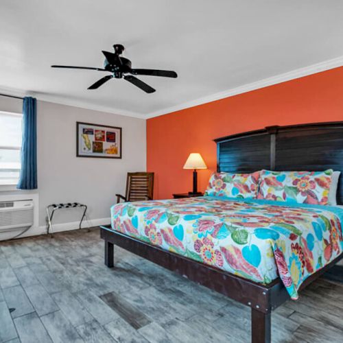 The image shows a bedroom with a colorful bedspread, two lamps, a ceiling fan, orange accent wall, and wooden flooring.