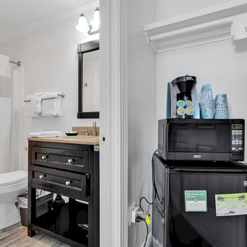 The image shows a bathroom with a toilet, vanity, and shower. Adjacent is a kitchenette with a microwave, coffee maker, and mini-fridge.