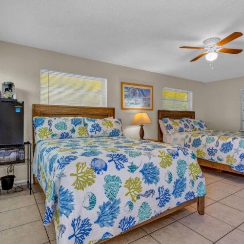 A bedroom with two double beds featuring floral bedding, a ceiling fan, framed art, a mini-fridge, and a coffee maker on a stand.