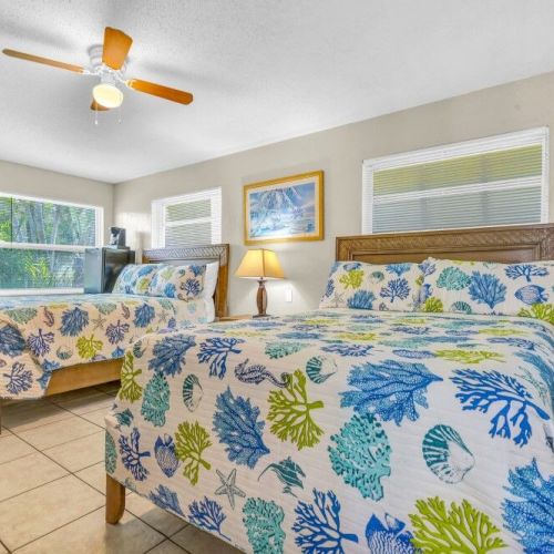 The image shows a bedroom with two beds covered in colorful, leaf-patterned bedspreads, a ceiling fan, a lamp, and artwork on the wall.