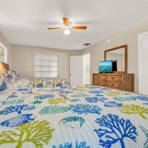 A bedroom with two beds featuring colorful ocean-themed bedding, a ceiling fan, window, dresser with a TV, and artwork on the walls.