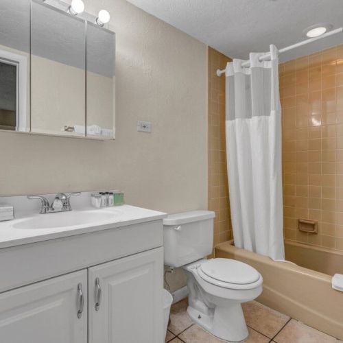 A clean bathroom with a sink, mirror, toilet, and a bathtub with a shower curtain. The tiles are beige, and there are towels and toiletries.