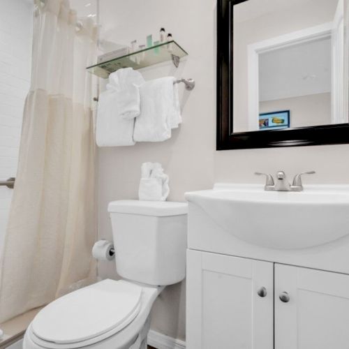 The image shows a bathroom with a white toilet, sink, mirror, towels, and shower with a curtain, all in a clean, minimalist design.