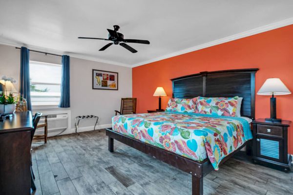 A bedroom with a colorful bedspread, two lamps, an orange accent wall, and a ceiling fan. There's a window and dresser visible.