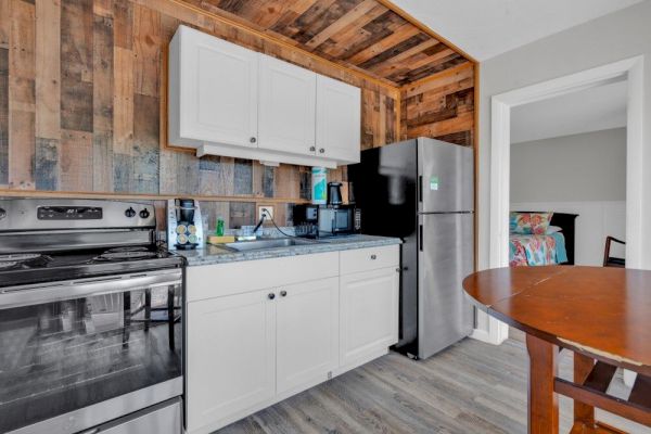 The image shows a cozy kitchen with wood paneling, a stove, a fridge, white cabinets, a round table, and a glimpse of a bedroom.