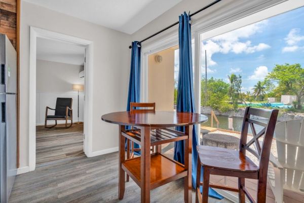 A cozy dining area with a round wooden table, chairs, blue curtains, and a view of a balcony with greenery outside under a blue sky.