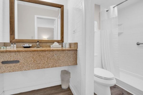 The image shows a modern bathroom with a large mirror, sink, toilet, shower, and wooden accents, featuring clean, neutral-colored decor.