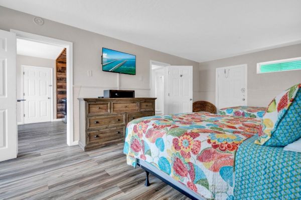 The image shows a bright bedroom with a colorful quilt on the bed, a dresser with a small TV, and an open door leading to another room.