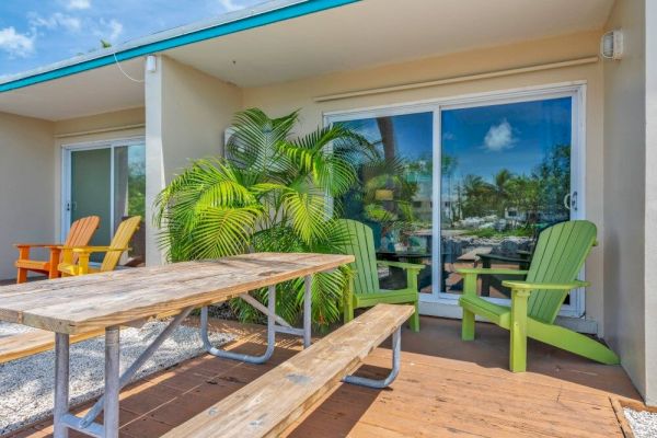 A patio with colorful Adirondack chairs, a wooden picnic table, lush plants, and a sliding glass door reflecting a sunny day.