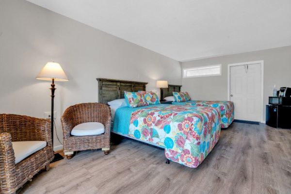 The image shows a bedroom with two beds having colorful bedding, wicker chairs, wooden flooring, and a lamp, creating a cozy atmosphere.