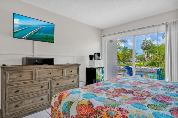 A colorful bedroom features a patterned bedspread, dresser, TV, and coffee maker; large windows reveal a pool and lush outdoor view.