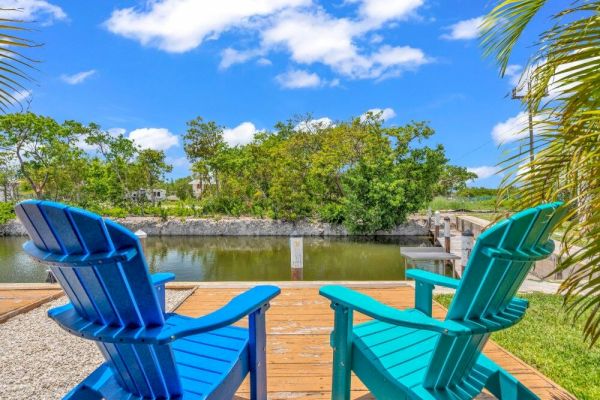 Two colorful chairs on a wooden deck overlook a tranquil canal surrounded by greenery, under a bright blue sky with fluffy clouds.