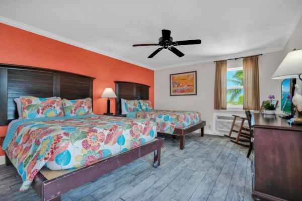 The image features a hotel room with two beds, colorful bedding, an orange accent wall, a ceiling fan, a window, and a TV on a desk.