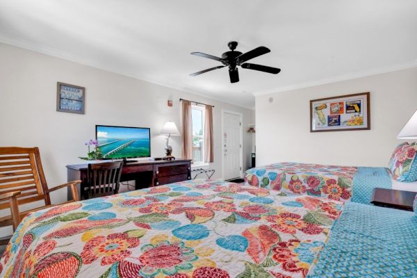 A brightly colored hotel room features two beds with floral quilts, a TV, ceiling fan, and a window with a view.