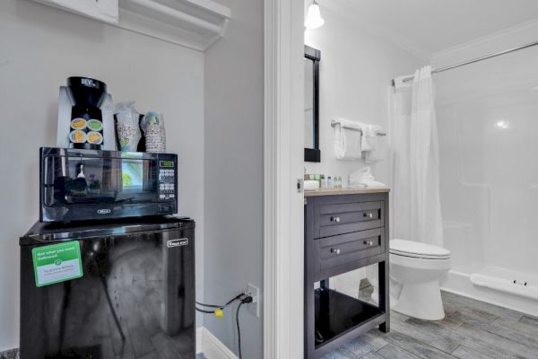 The image shows a kitchenette with a coffee maker, microwave, and mini-fridge next to a bathroom with a vanity, toilet, and shower.