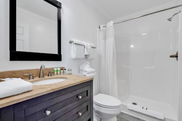 A modern bathroom with a black-framed mirror, wooden vanity, toilet, and white shower curtain around a standing shower.